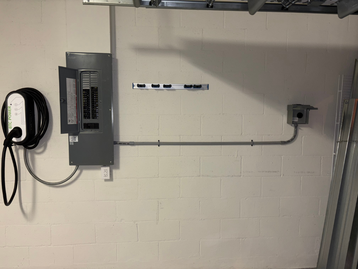 An electric vehicle charging station and electrical panel are mounted on a white cinder block wall in a garage.