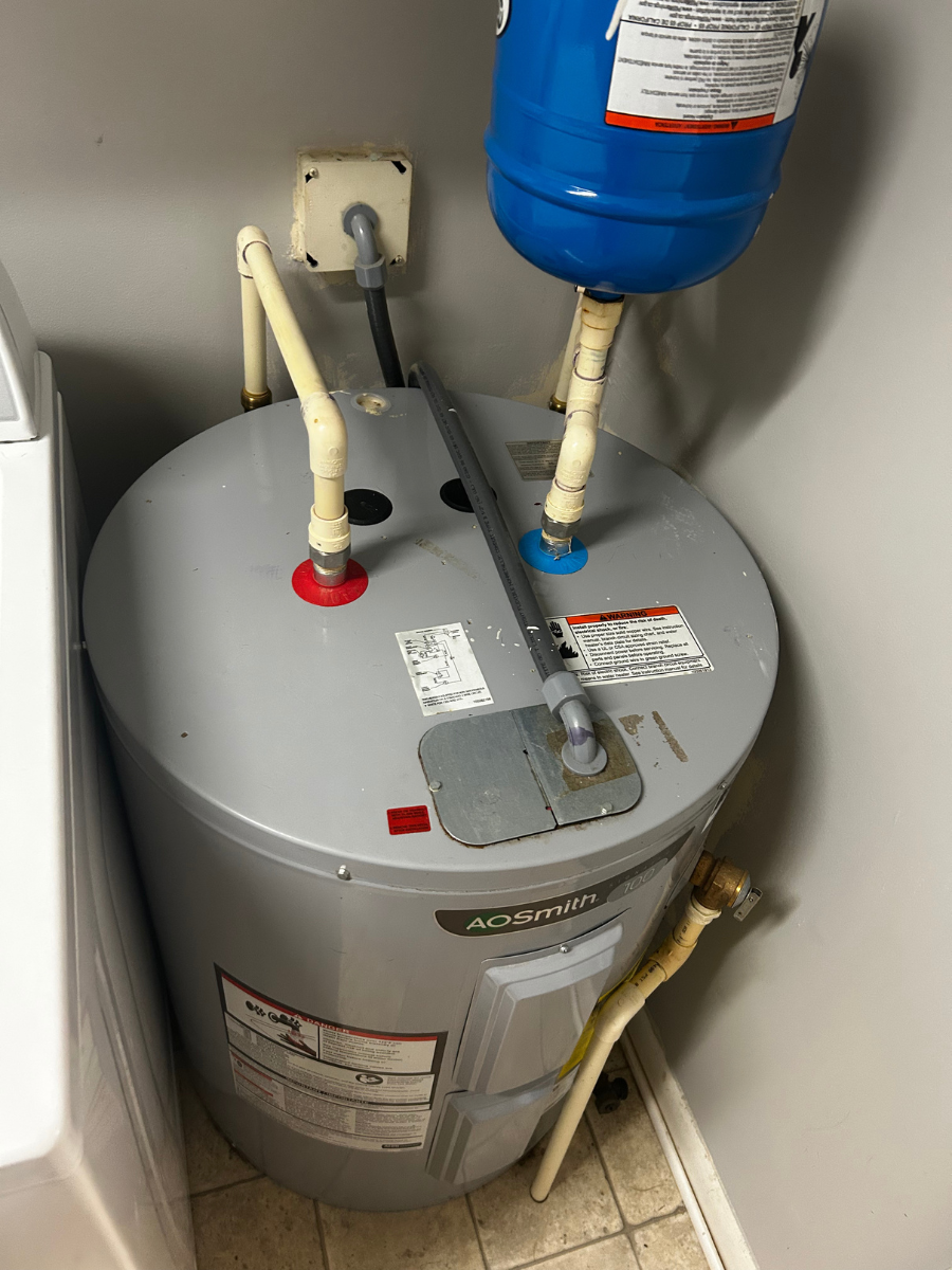 An electric water heater with a blue expansion tank on top, connected by white pipes in a corner near a white appliance.