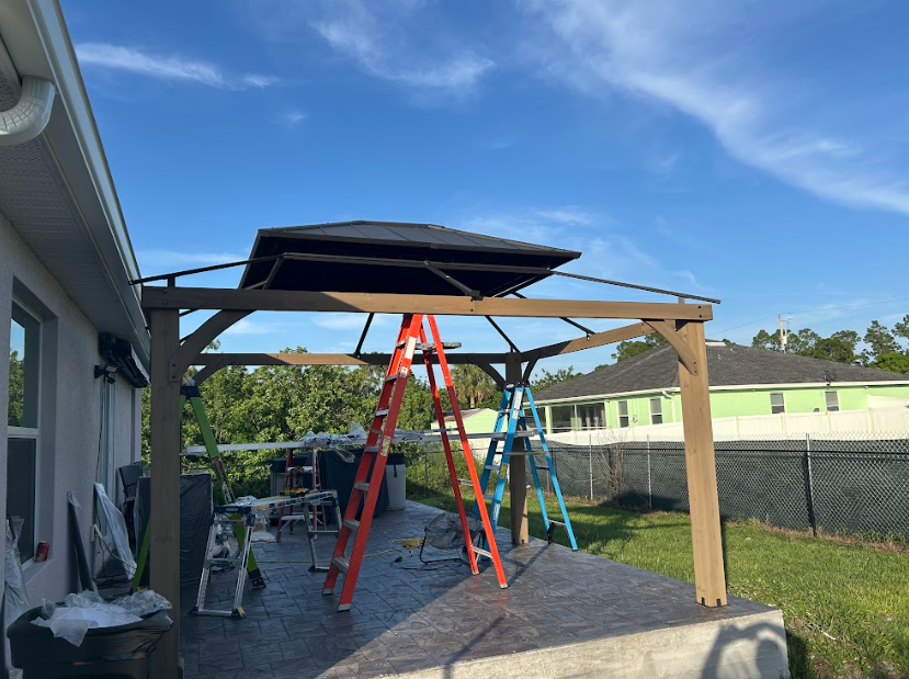 Gazebo under construction on a concrete patio, with two ladders and blue sky.