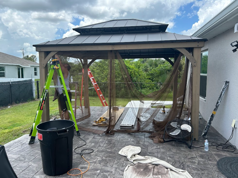 Gazebo being assembled on a patio. Brown wood frame, dark roof, mesh netting, tools scattered around.