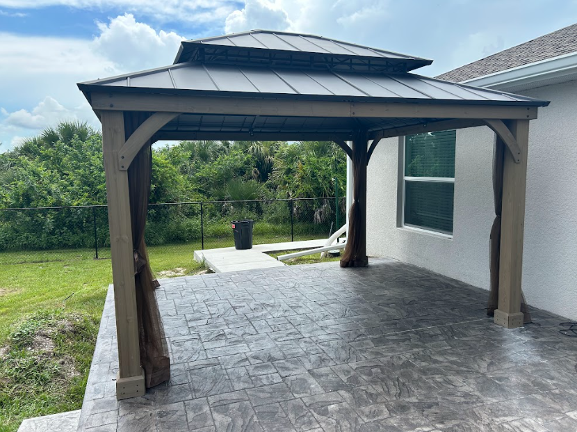 Gazebo with brown supports and metal roof on a concrete patio, next to a house with a green lawn.