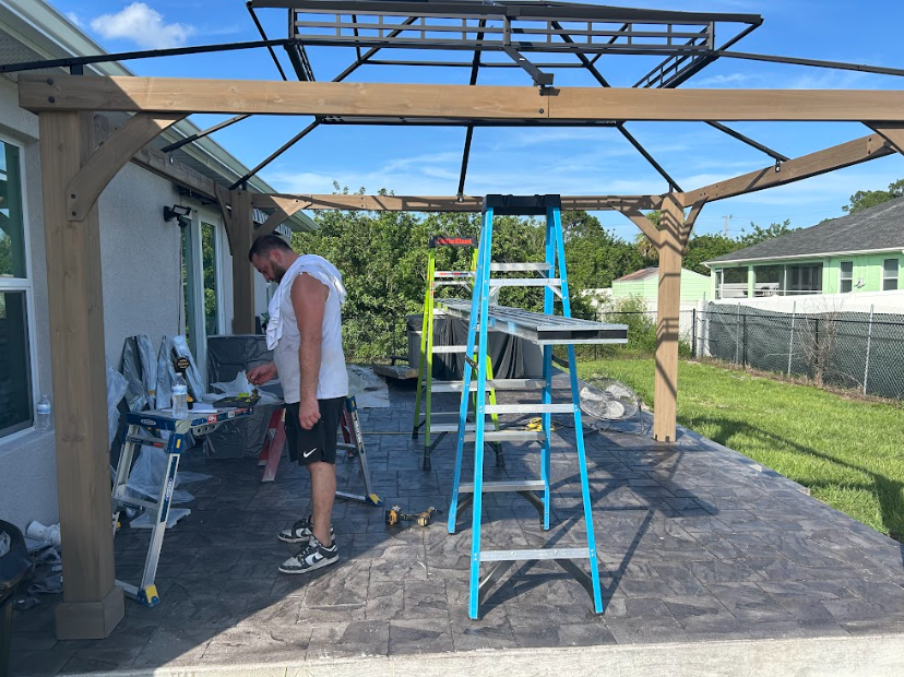 Man working on gazebo frame on a patio. Green ladder, blue sky, and lush green grass.