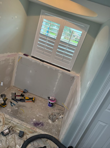Bathroom renovation in progress, drywall, tools, window with shutters. Light blue walls and white trim.