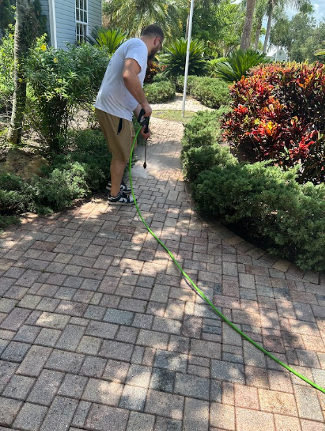 Man spraying a brick pathway with a green hose in a garden.