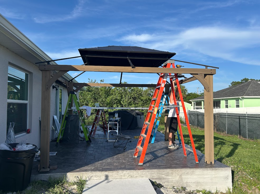 Man on ladder installing gazebo on patio with blue sky.