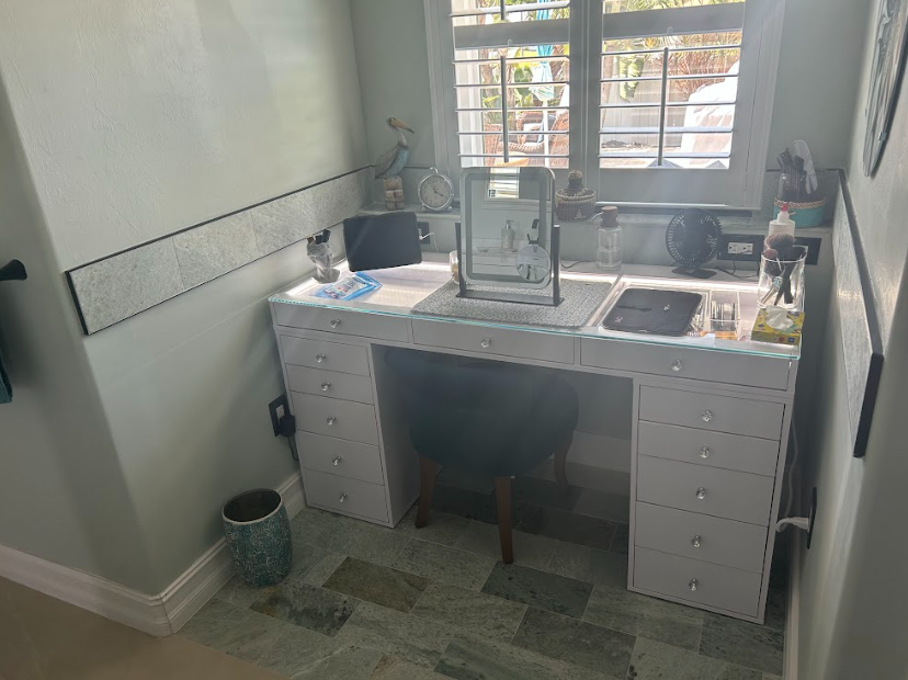 White vanity table with drawers in a bathroom, black chair, window, and accessories.