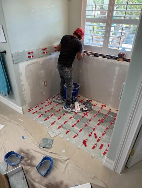 Man tiling a bathroom; gray tiles, red spacers, window, blue bucket, light blue walls.