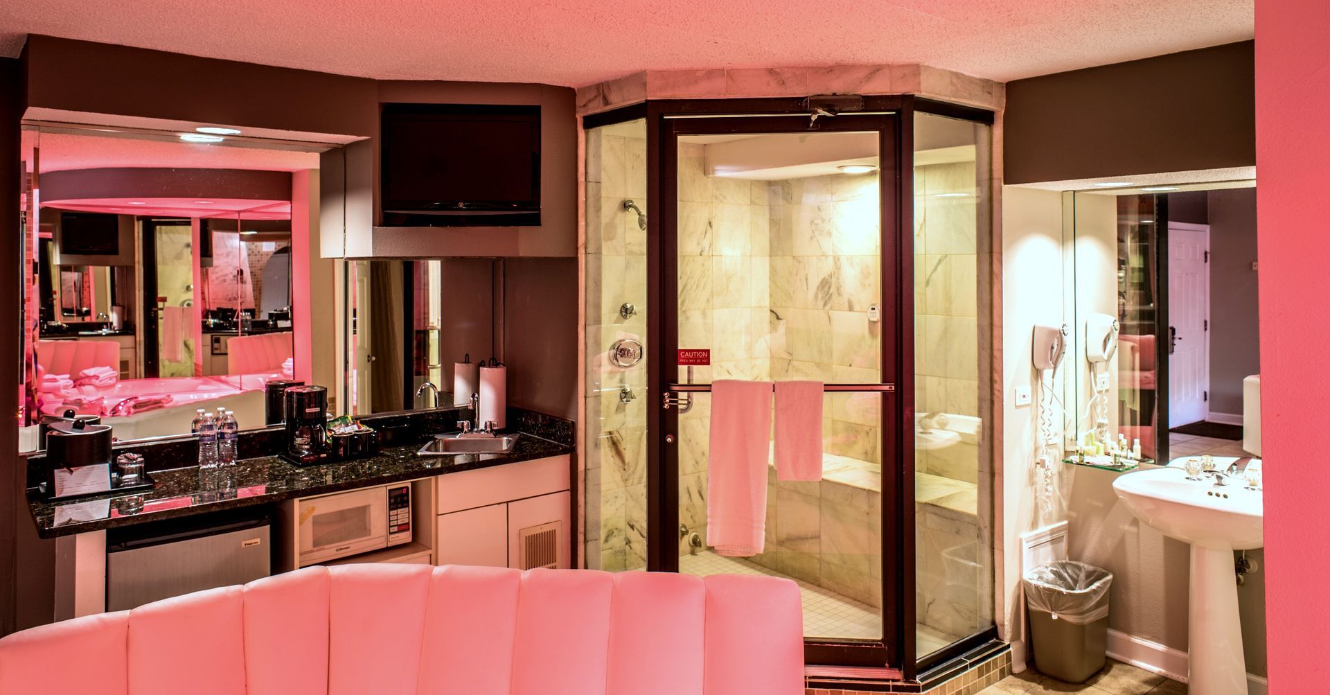 A motel room with a pink sofa, kitchenette, and a glass-walled shower. Soft pink and gray tones.