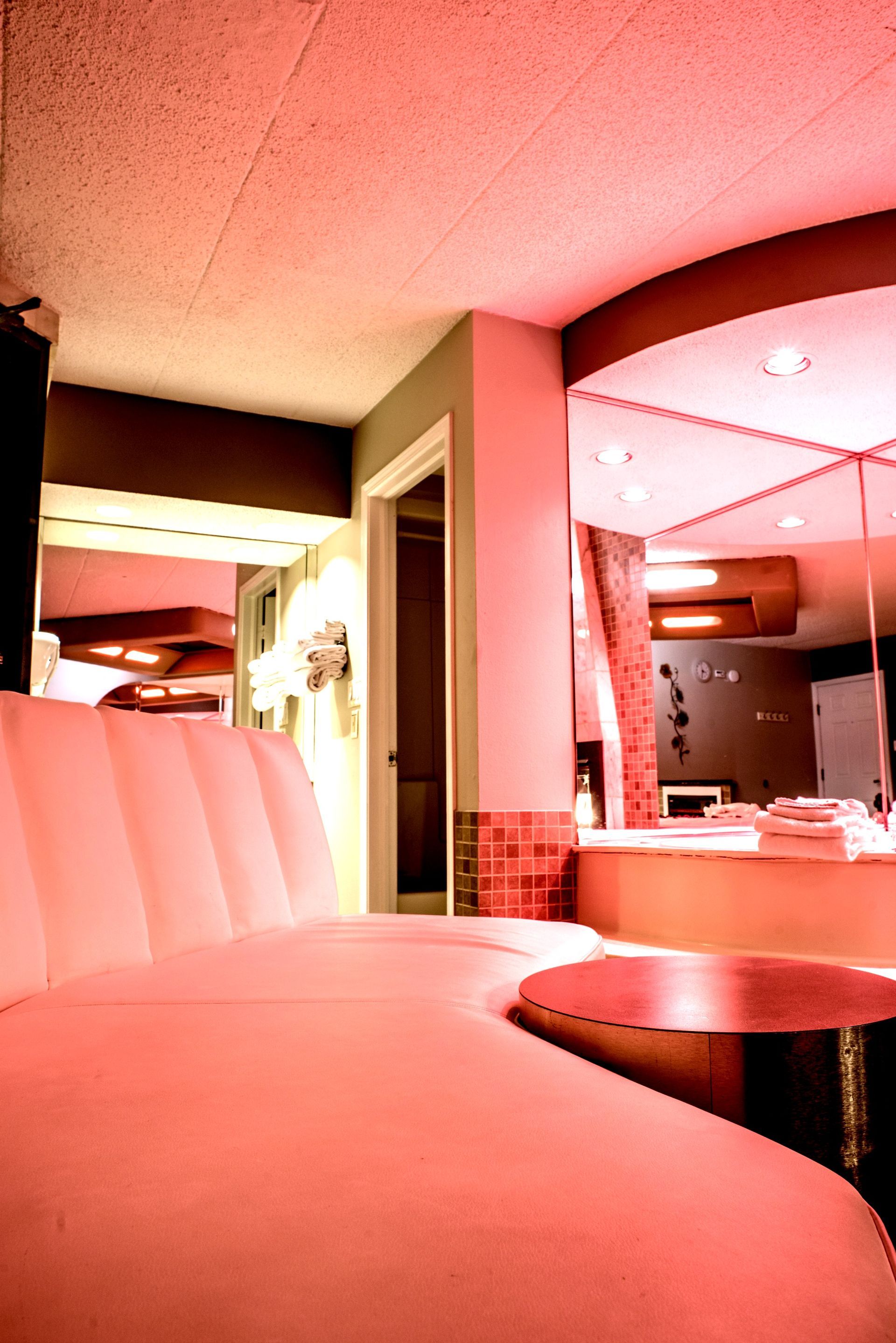 Pink-lit curved sofa in a room with mirrors and a small table; retro aesthetic.
