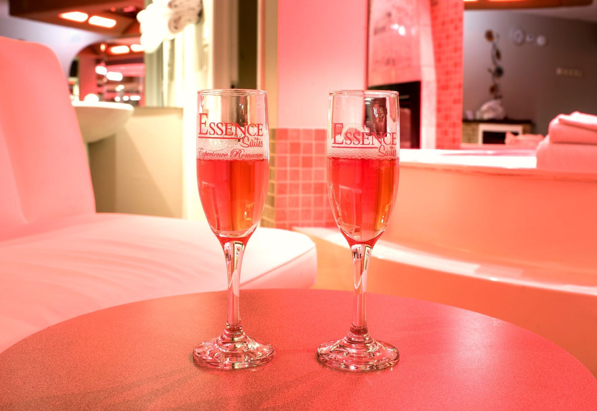 Two champagne flutes filled with pink liquid sit on a table, with a pink-lit interior in the background.