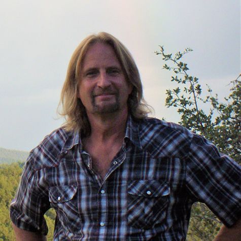 James Lazok with long blonde hair and a goatee wearing a plaid shirt, standing outdoors against a hillside background.