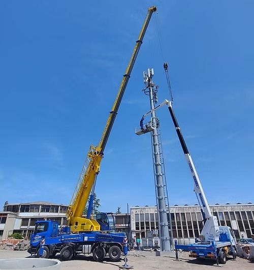 Due gru sollevano attrezzature su una torre per cellulari in costruzione contro un cielo azzurro.