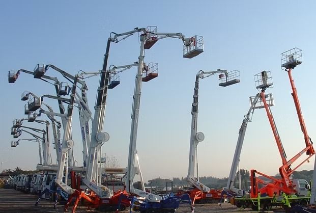 Piattaforme aeree parcheggiate in un parcheggio, vari modelli con bracci e piattaforme estesi.