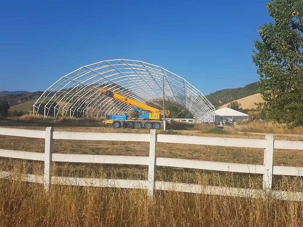 Cantiere con una grande struttura metallica ad arco sotto un cielo azzurro e limpido, su cui lavora una gru gialla.