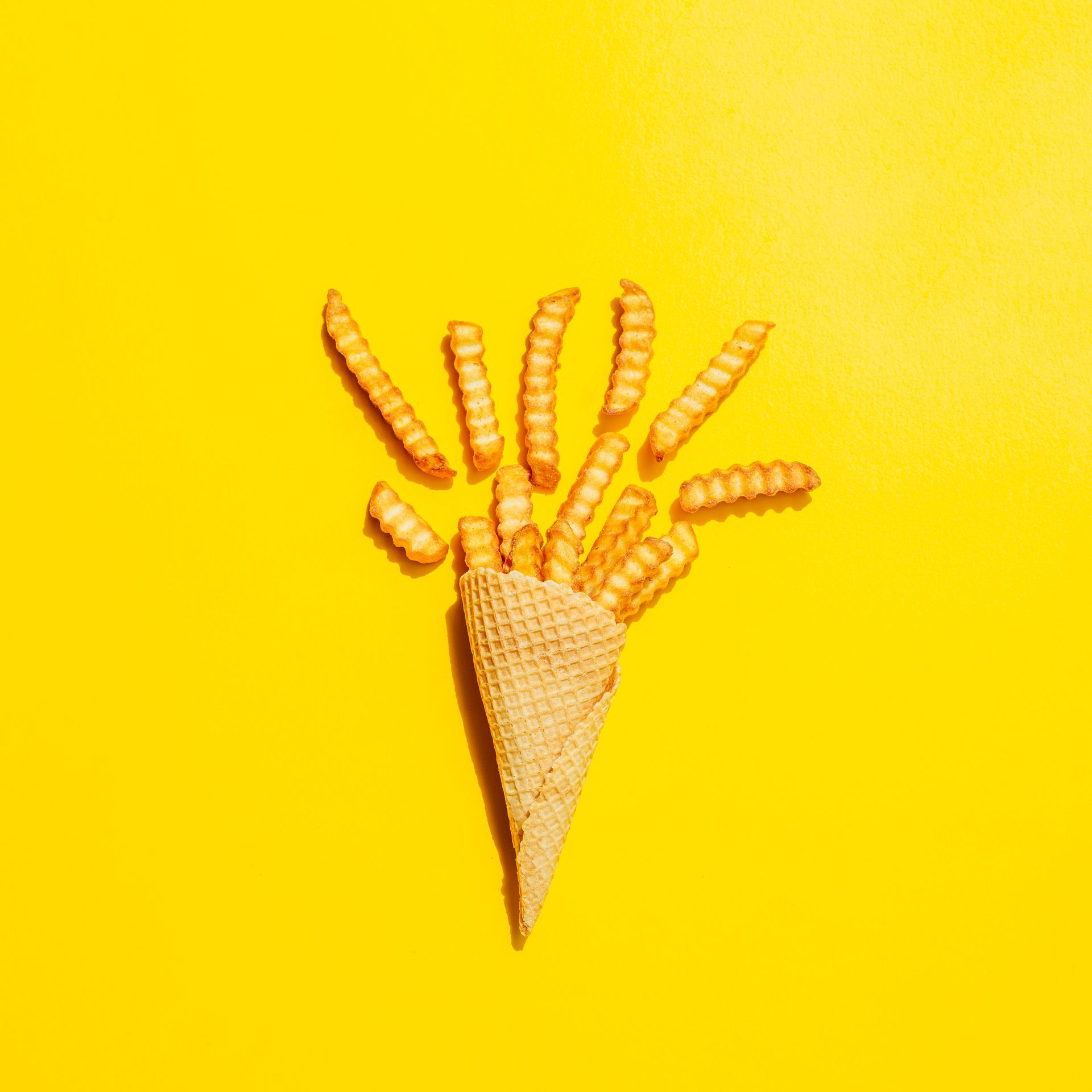 Fond jaune. Un cornet gaufré garni de frites ondulées, dont certaines débordent.