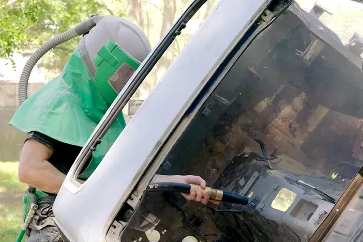 A man wearing a green helmet is sandblasting a car.