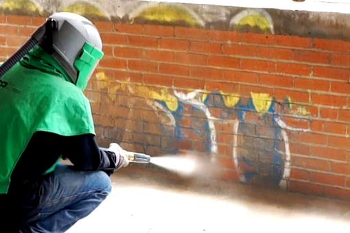 A man is sandblasting graffiti on a brick wall.