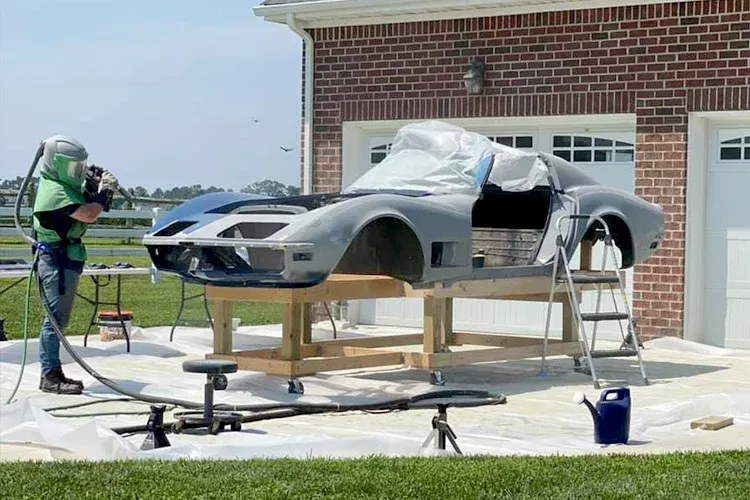 A man is sandblasting a car in front of a brick house.
