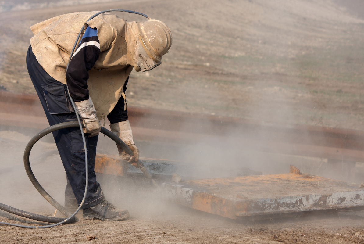 A man is sandblasting a piece of metal in the dirt.