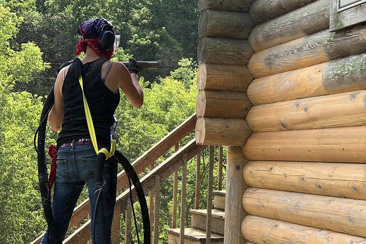 A man is sandblasting a log cabin with the blasted wood looking brand new and an obvious dividing line from the uncleaned wood
