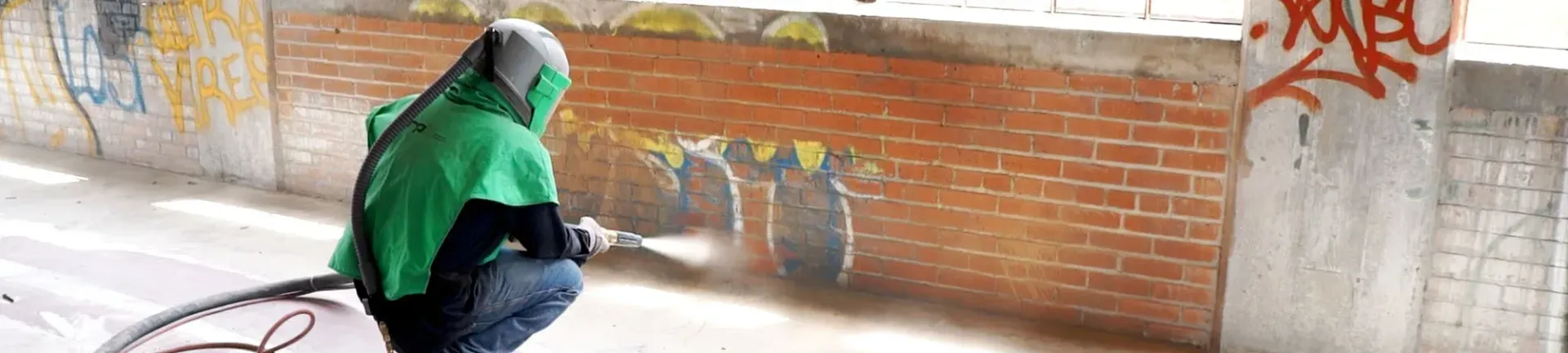 A person in a green hoodie is kneeling down in front of a brick wall with graffiti on it.