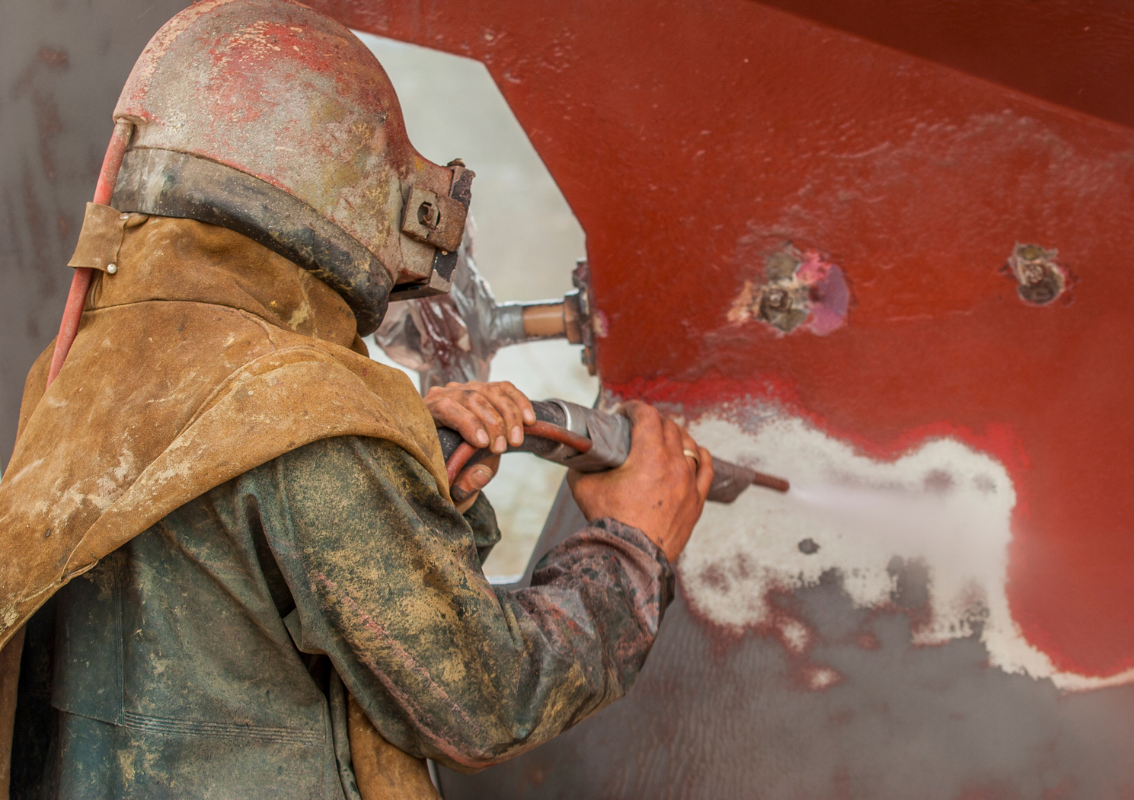 A man wearing a helmet is sandblasting a boat.