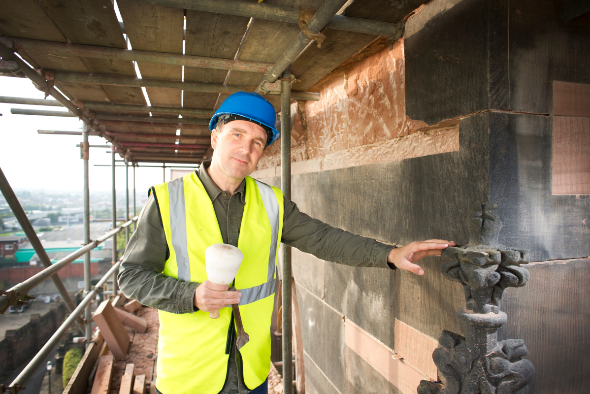 A man wearing a hard hat and a yellow vest is standing on a scaffolding.