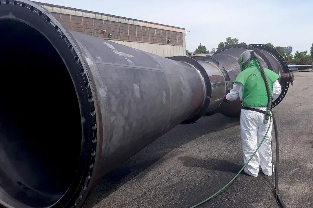 A man in a green suit is sandblasting a large pipe.
