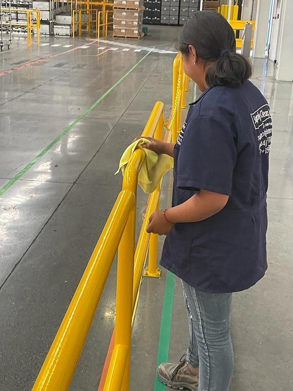 Una persona con uniforme azul limpiando una barandilla de seguridad amarilla en un almacén.