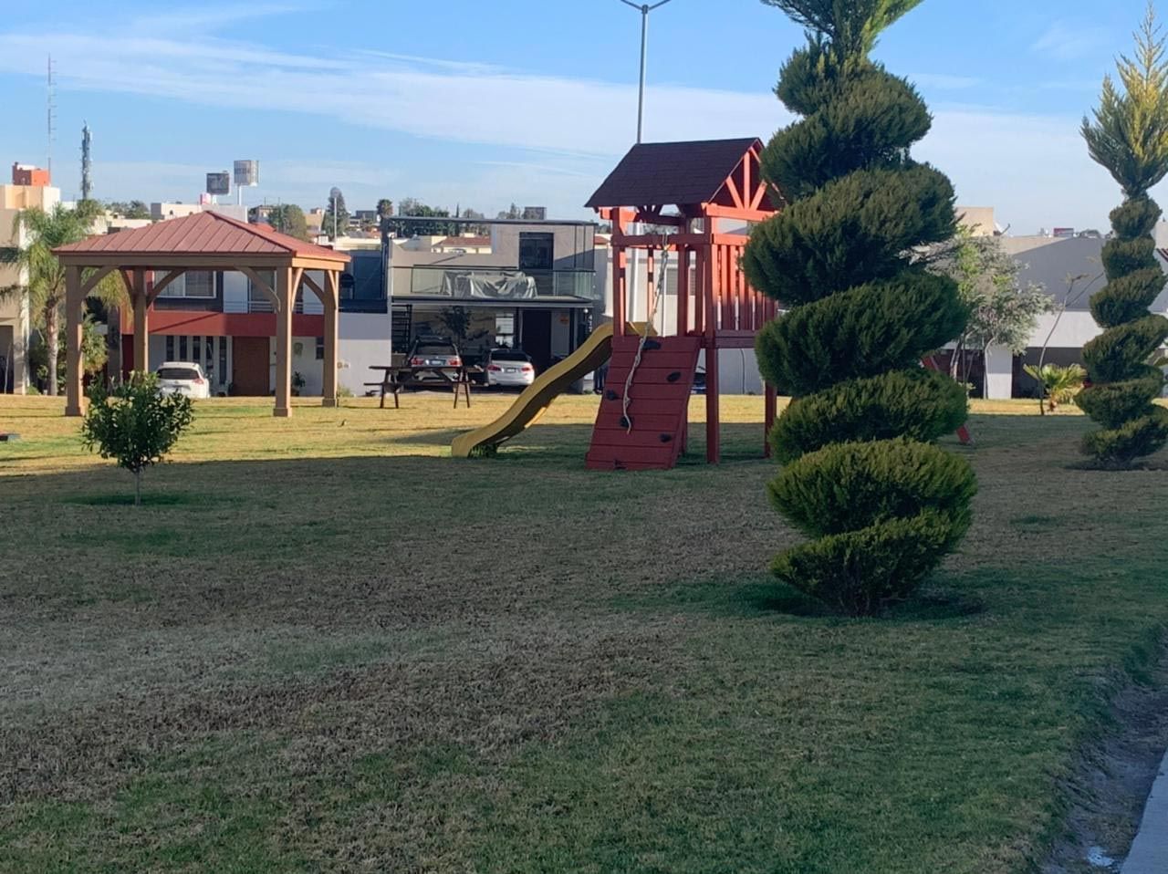 Parque con zona de juegos y arbustos decorativos en espiral. Se ven edificios al fondo.