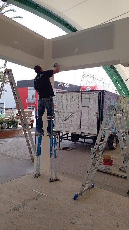 Hombre en zancos trabajando en un techo alto. Obra con escaleras y remolque al fondo.