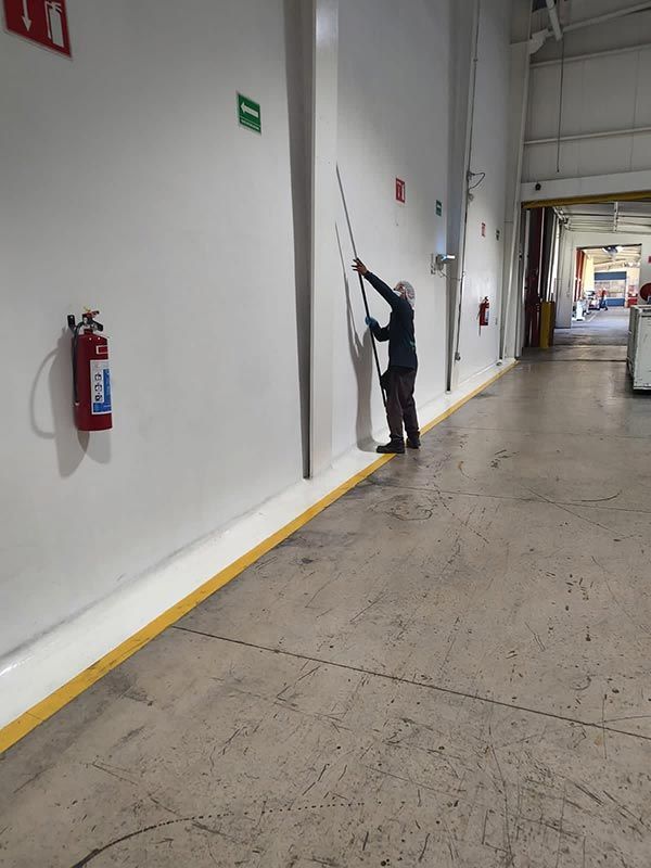 Persona que usa un palo para alcanzar algo en una pared blanca alta dentro de un edificio industrial.