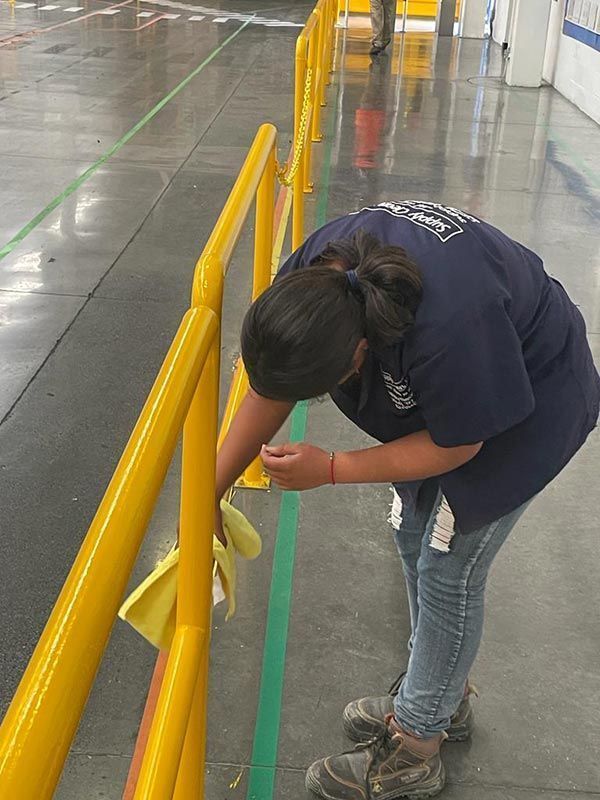 Una mujer con camisa azul limpia un pasamanos amarillo con un paño amarillo en un entorno industrial.