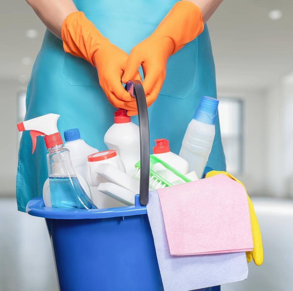 Una persona con guantes naranjas y delantal azul sostiene un balde azul lleno de productos de limpieza.