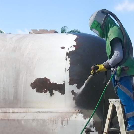 A man wearing a green vest is sandblasting a tank