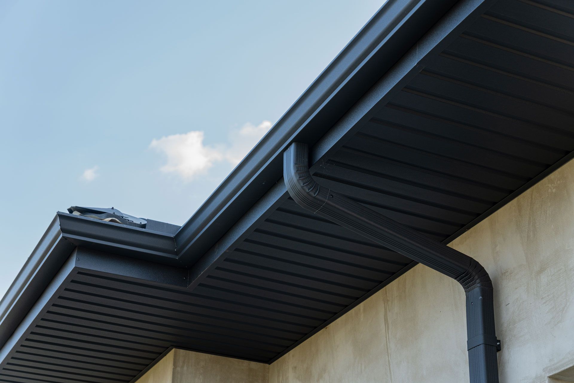 Black rain gutter and downspout on a beige wall under a dark roof and a blue sky.