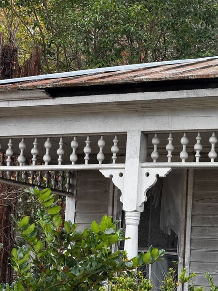White porch with ornate railing and a weathered roof; overgrown foliage