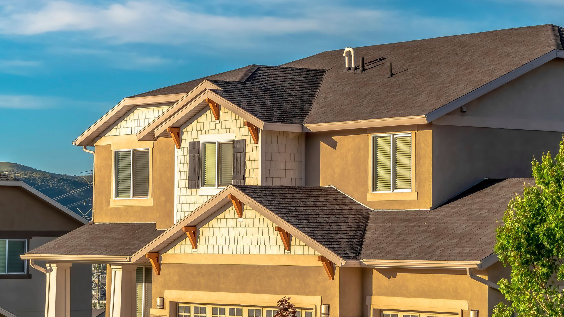 Two-story house with tan stucco exterior, dark roof, and blue sky background.