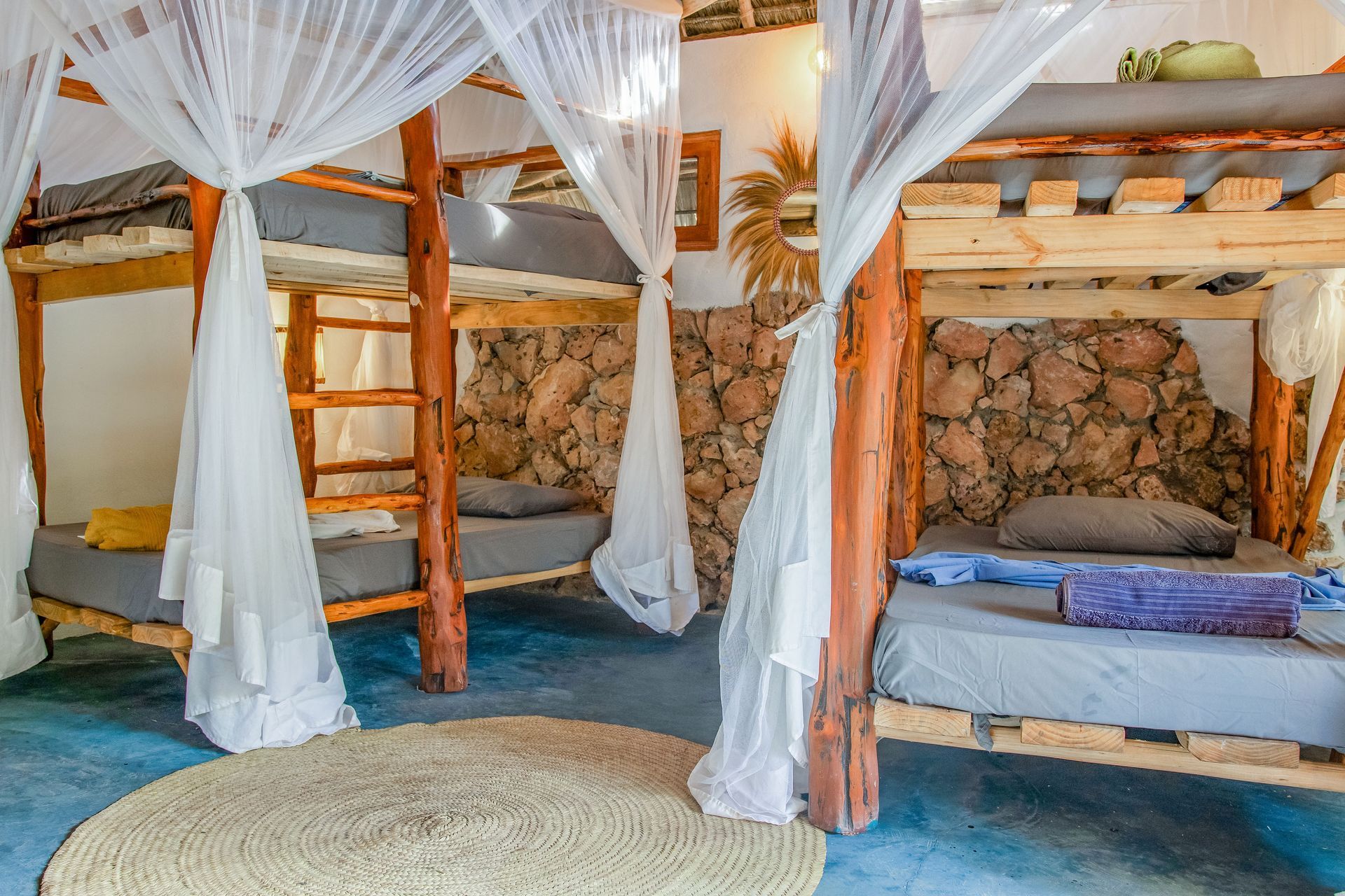 Bunk beds in a brightly lit room. Each bed has a white mosquito net. Stone walls. A beige rug.