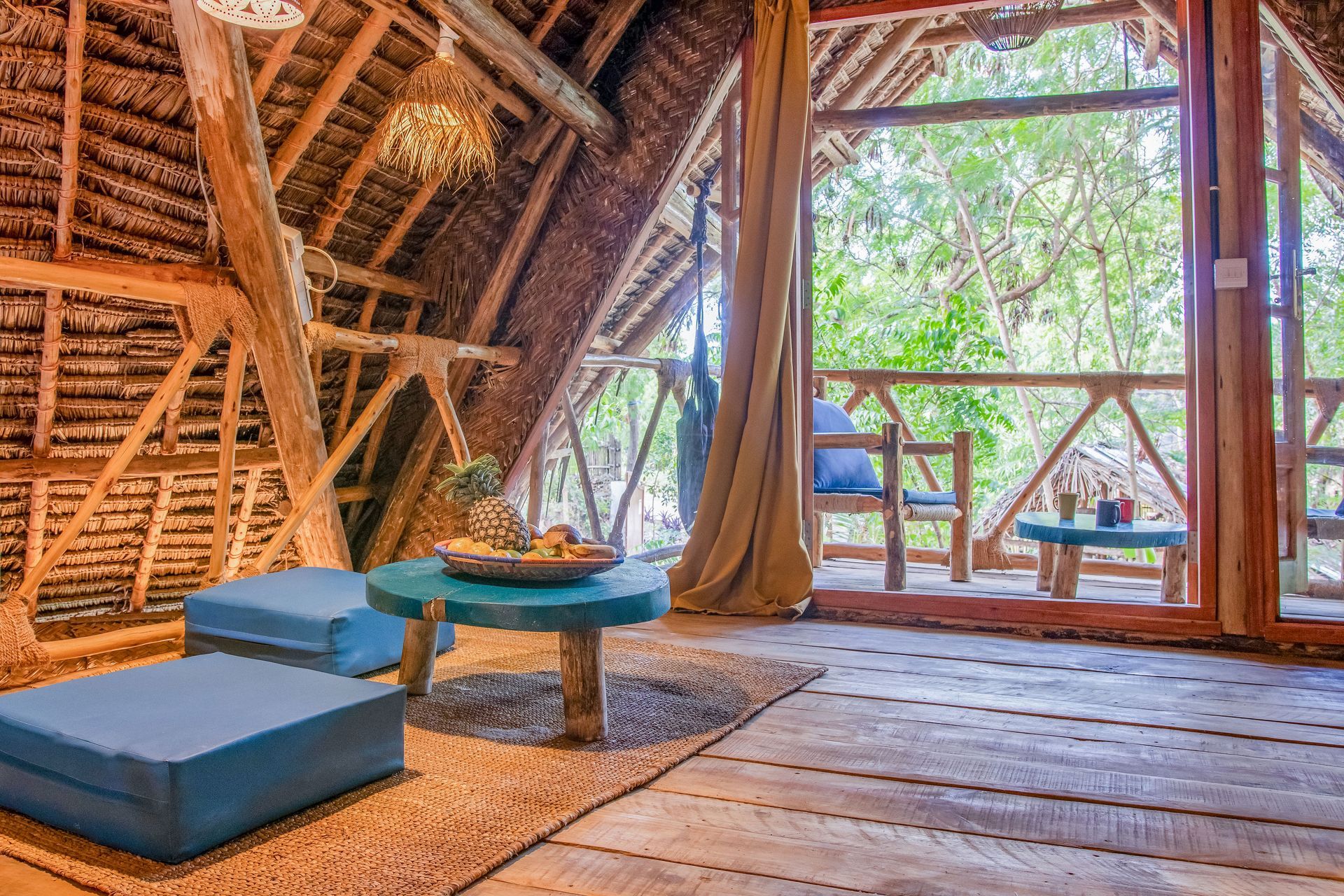 Cozy treehouse interior with blue cushions, turquoise table, and balcony overlooking lush greenery.