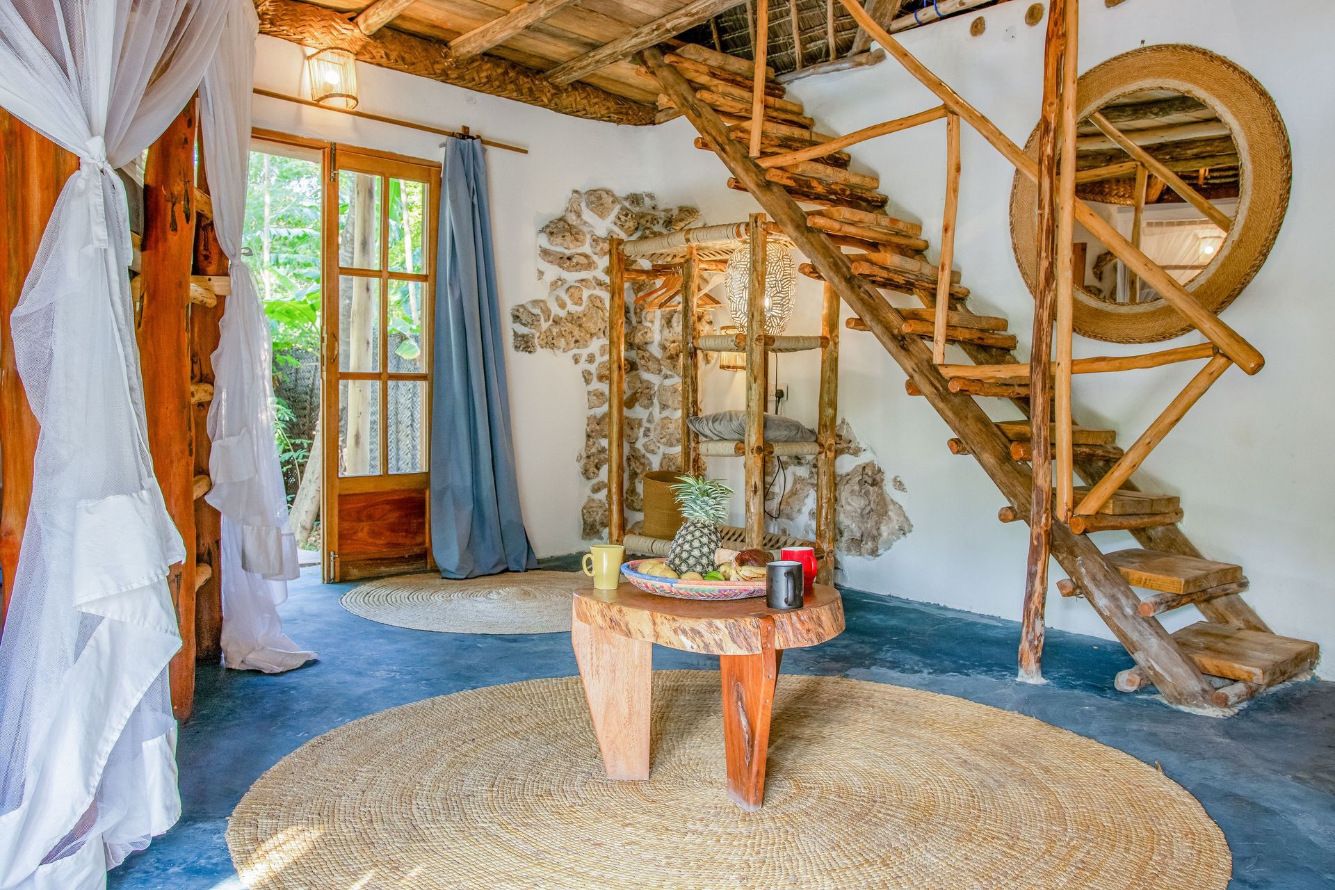 Rustic interior with wooden staircase, circular rug, stone wall, and open door to nature.