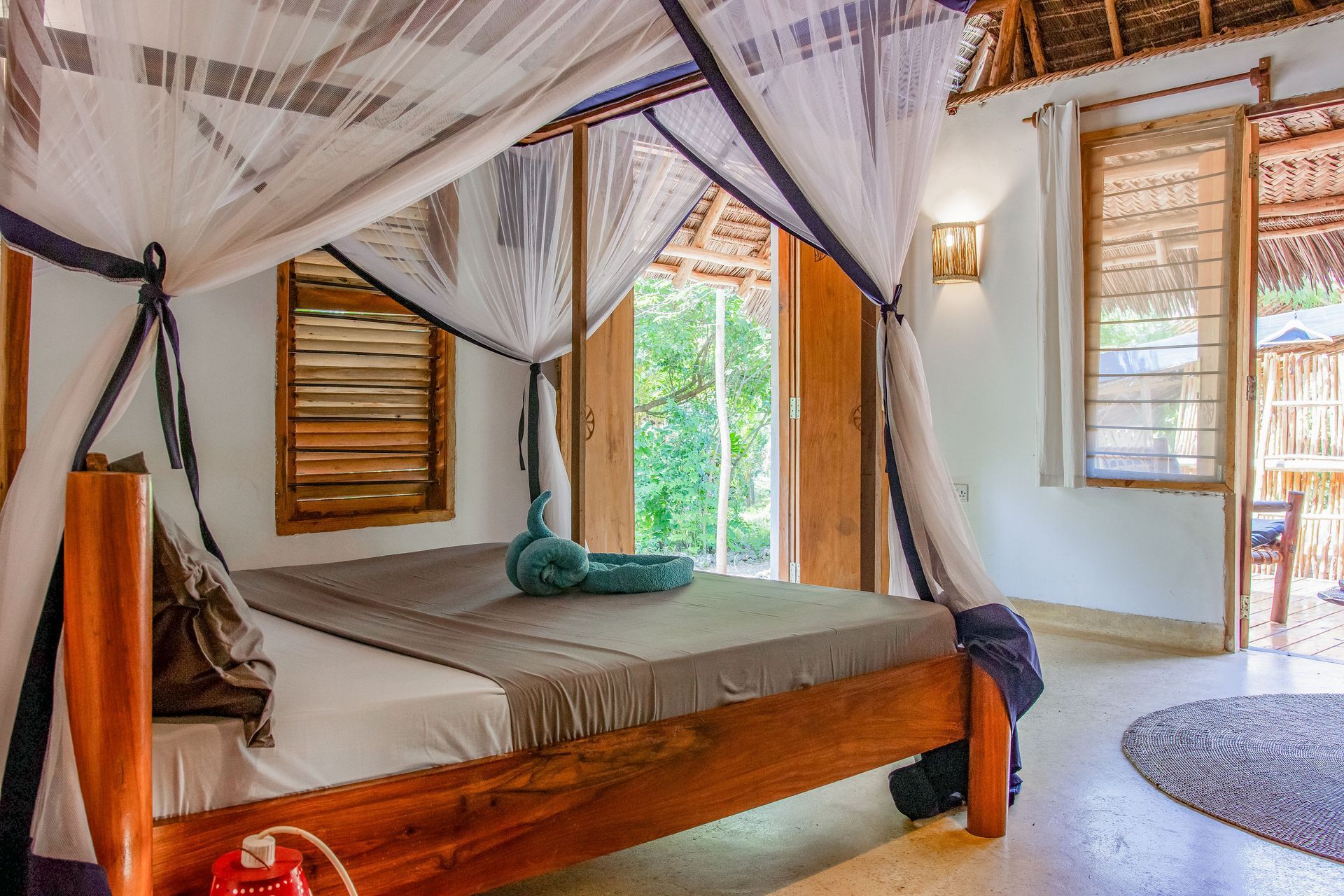 Bedroom with wooden bed frame, mosquito net, open doors to a tropical view.