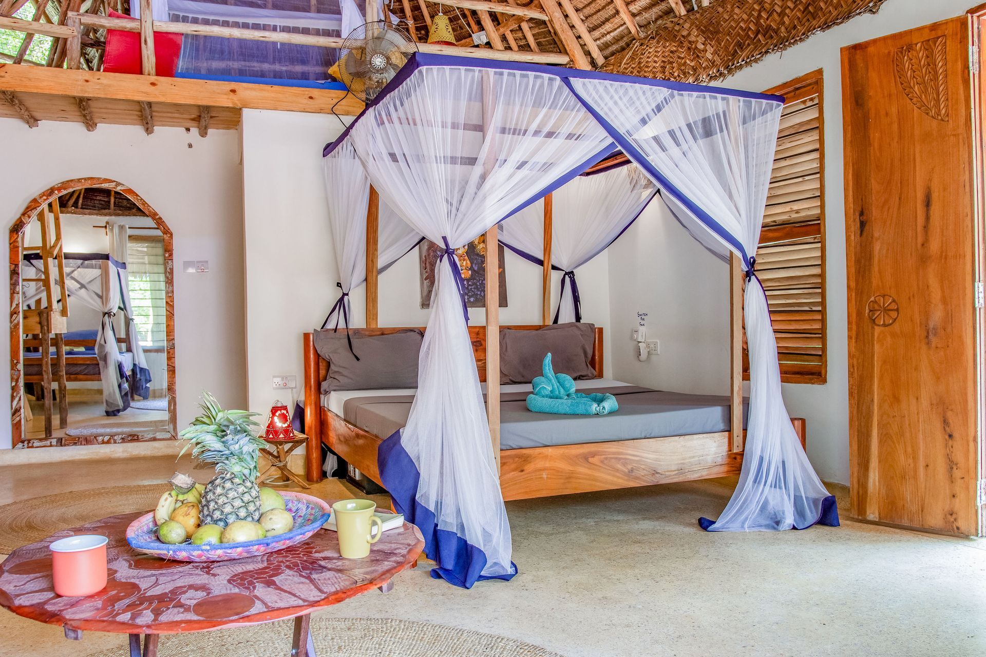 Bedroom with canopy bed, fruit platter, and open door. White and blue mosquito net over wooden bed.