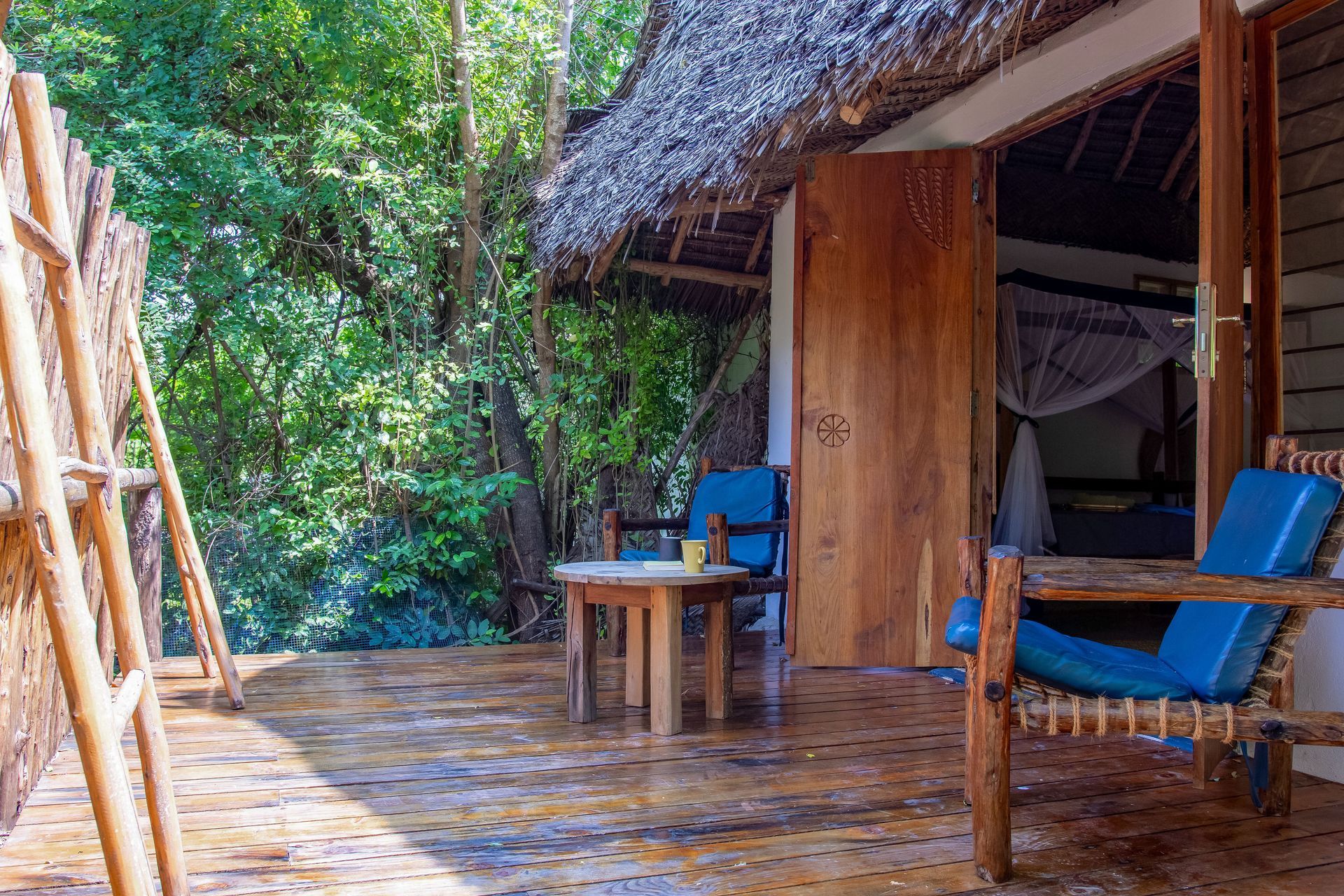 Patio with blue chairs, small table, and a thatched roof, opening to a bedroom with mosquito net.