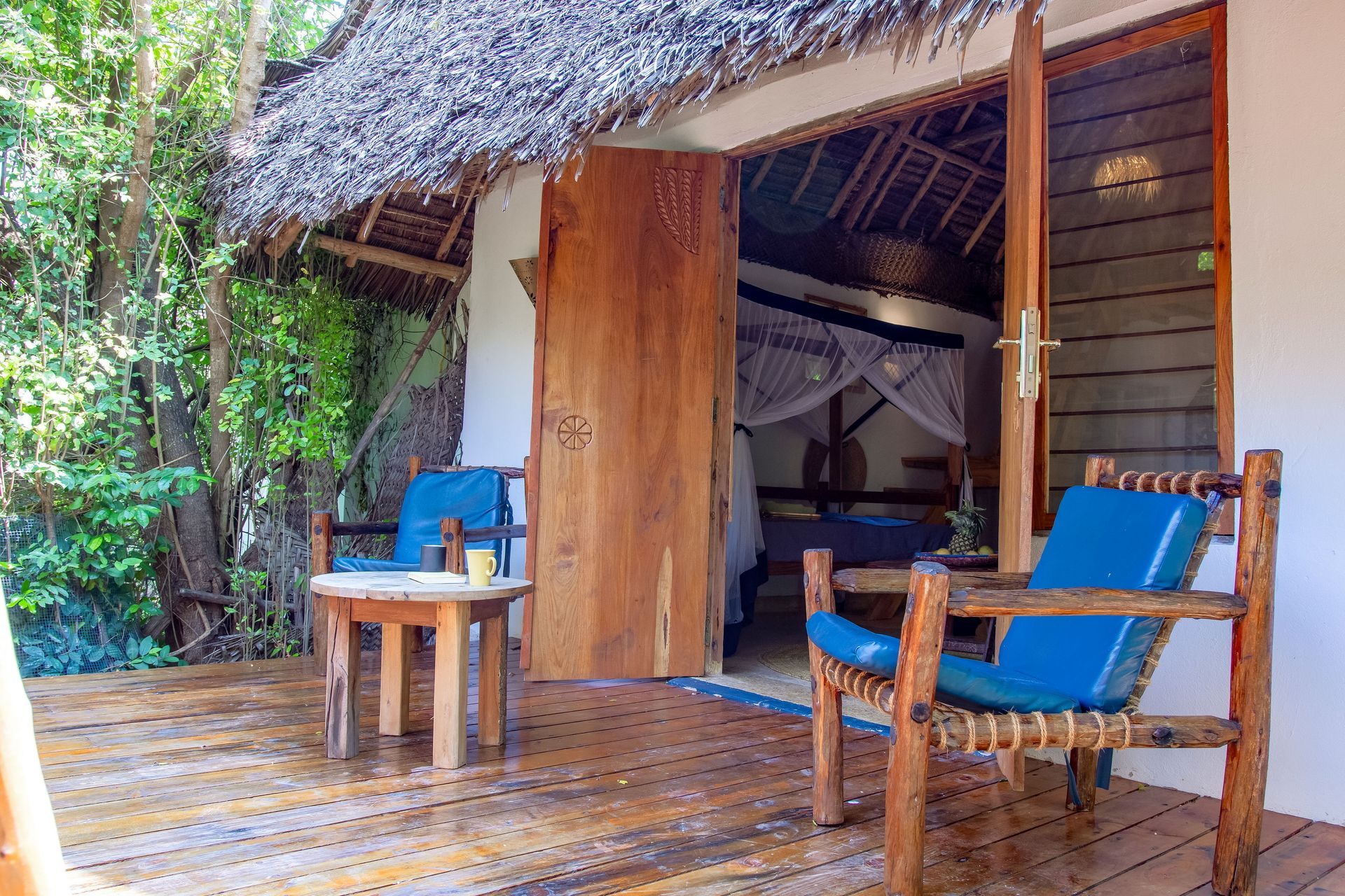 Wooden deck with blue chairs, a small table, and open doorway to a bedroom in a tropical setting.