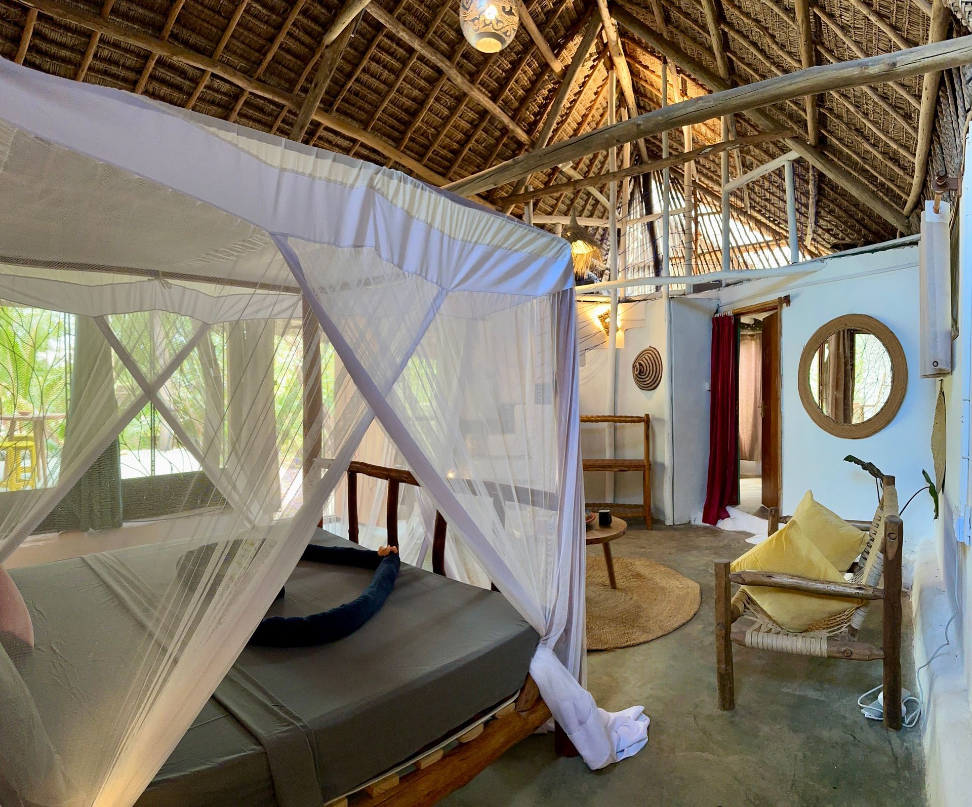 Bedroom with mosquito net over a bed, thatched roof, and a circular mirror on the wall.