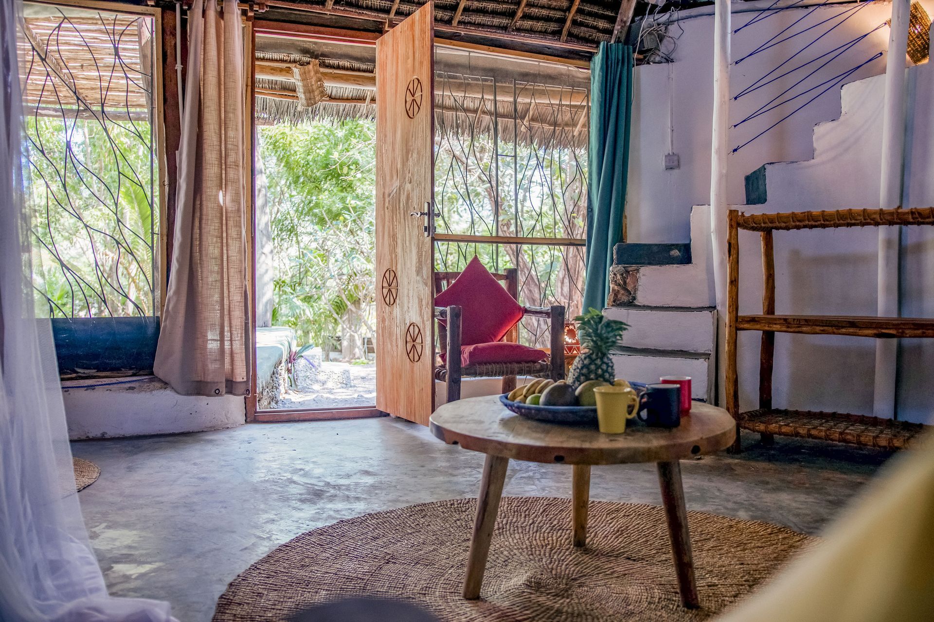 Cozy living room with open doors, a round table, fruit, and a view of lush greenery.