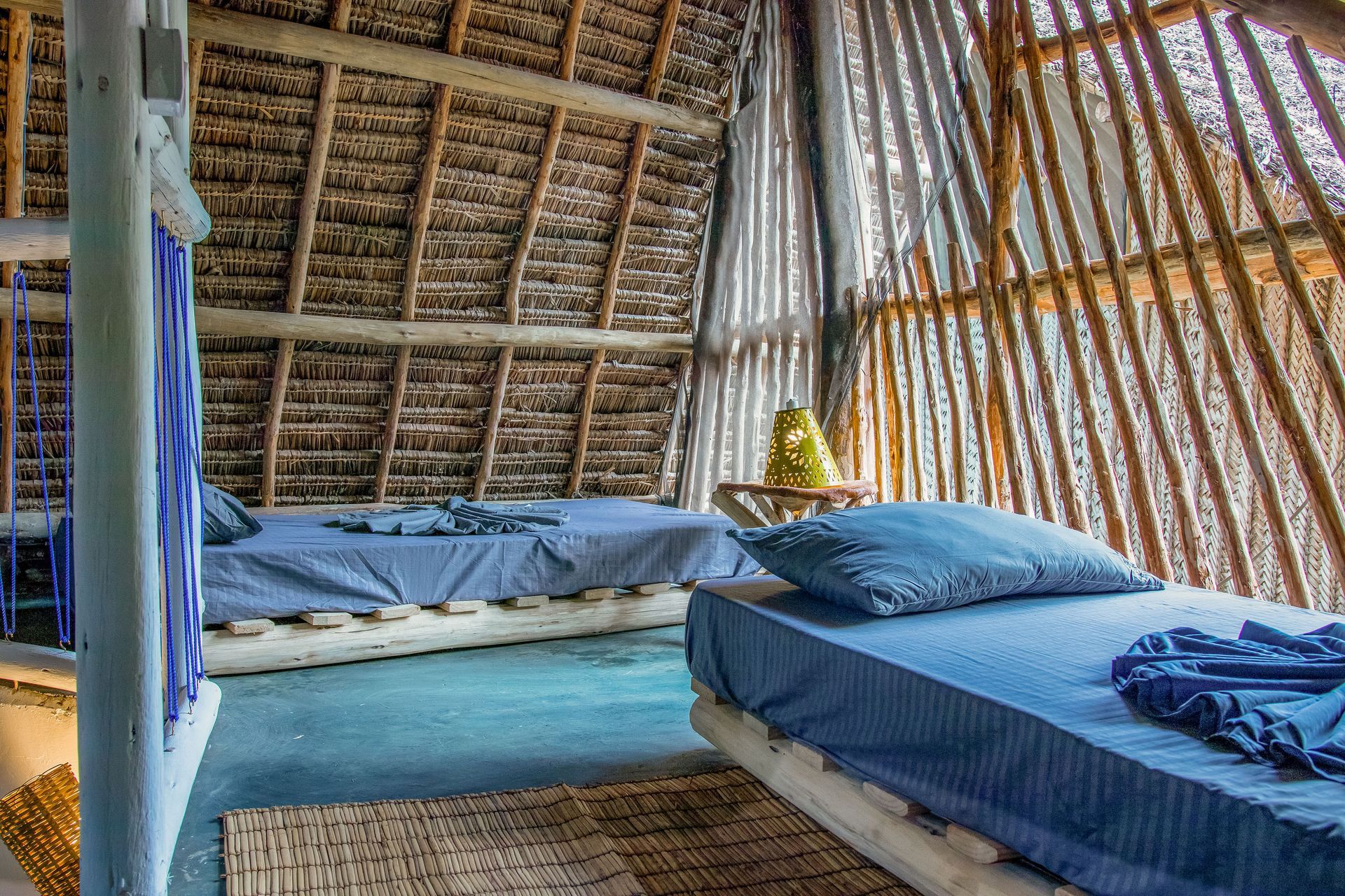 Two beds in a rustic room with a thatched roof and bamboo walls. Blue bedding, natural light.