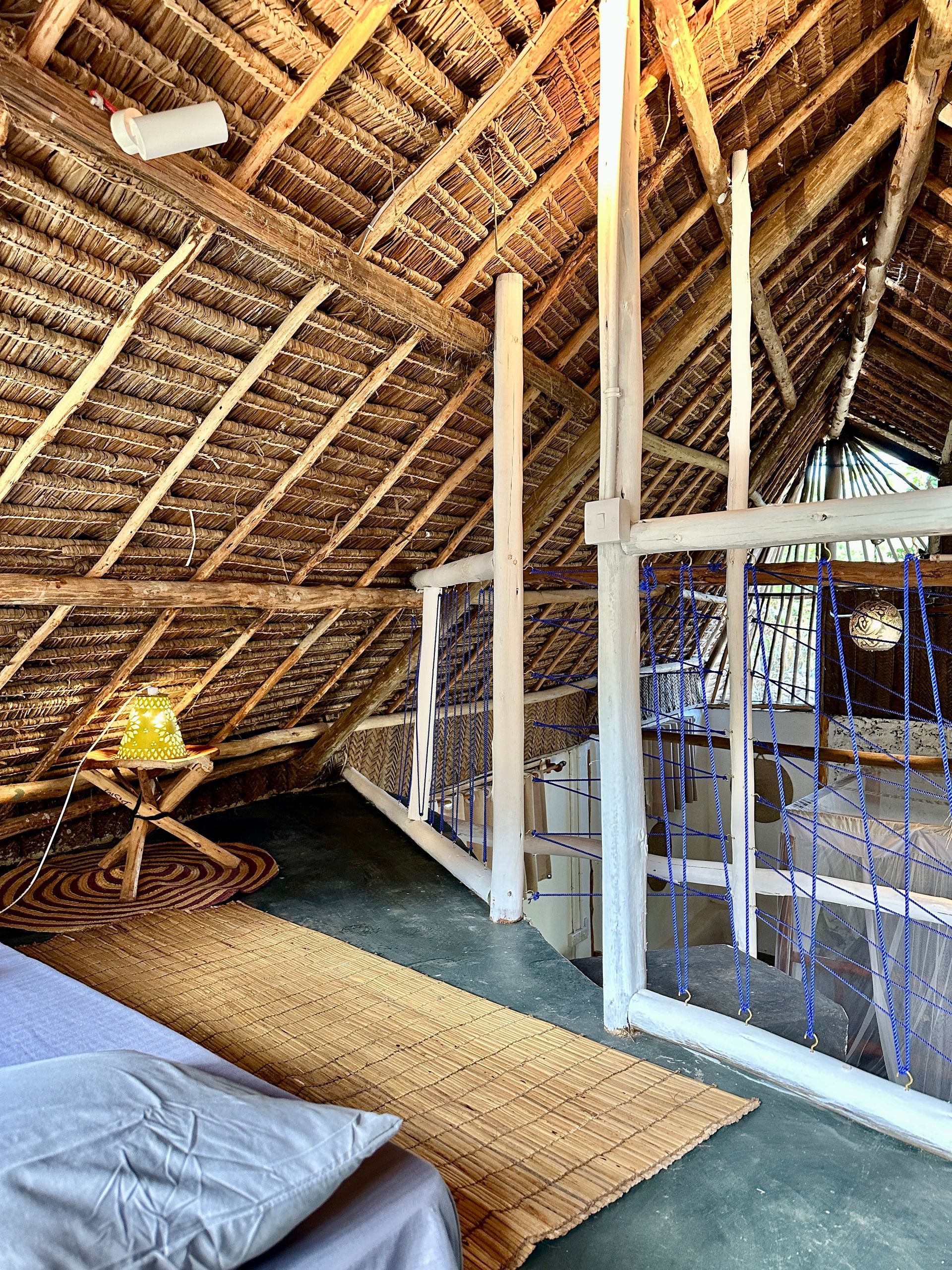 Loft bedroom with thatched roof, bed, rug, lamp, and white posts.