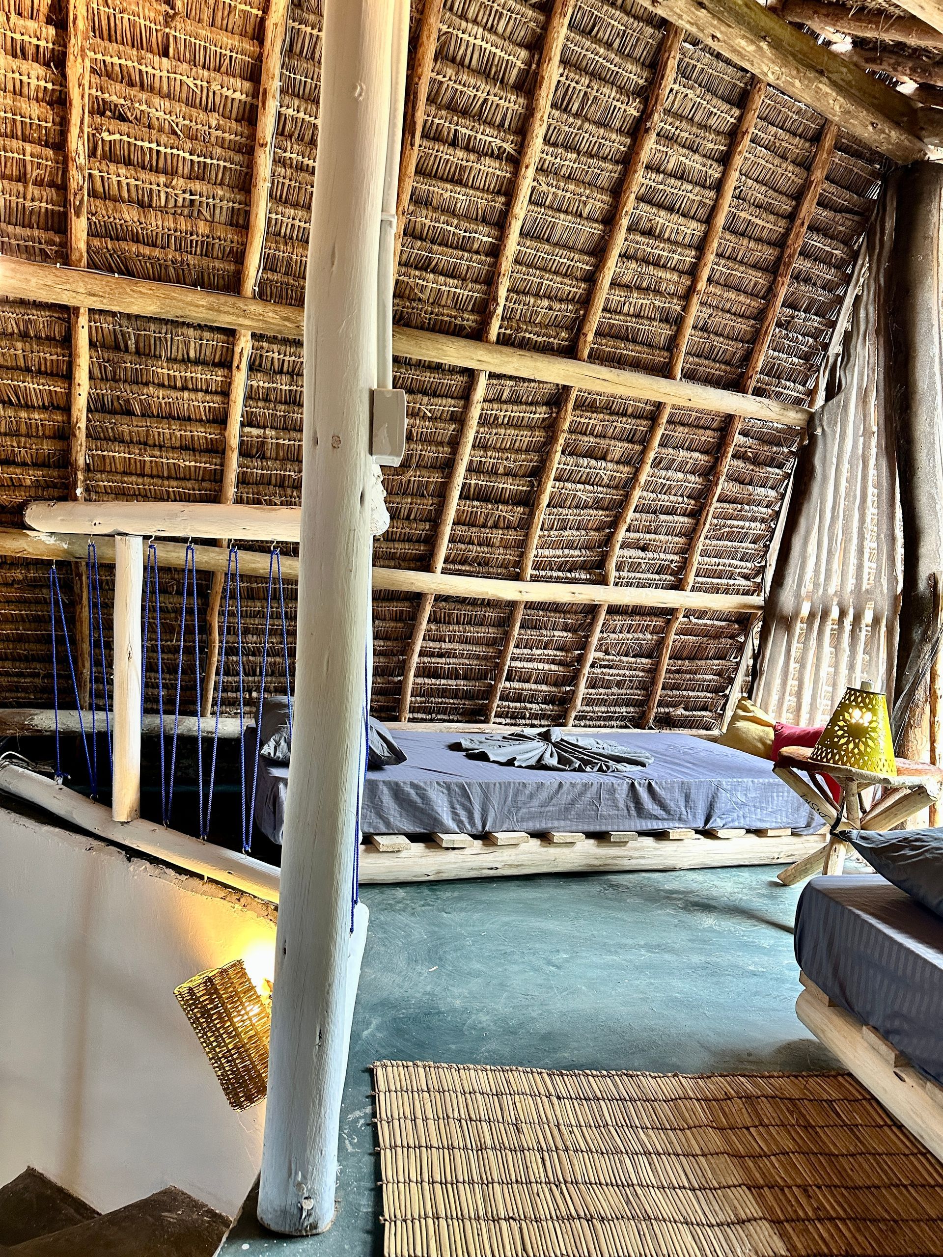 Bedroom with thatched roof, wooden frame bed, blue bedding, and woven rug.